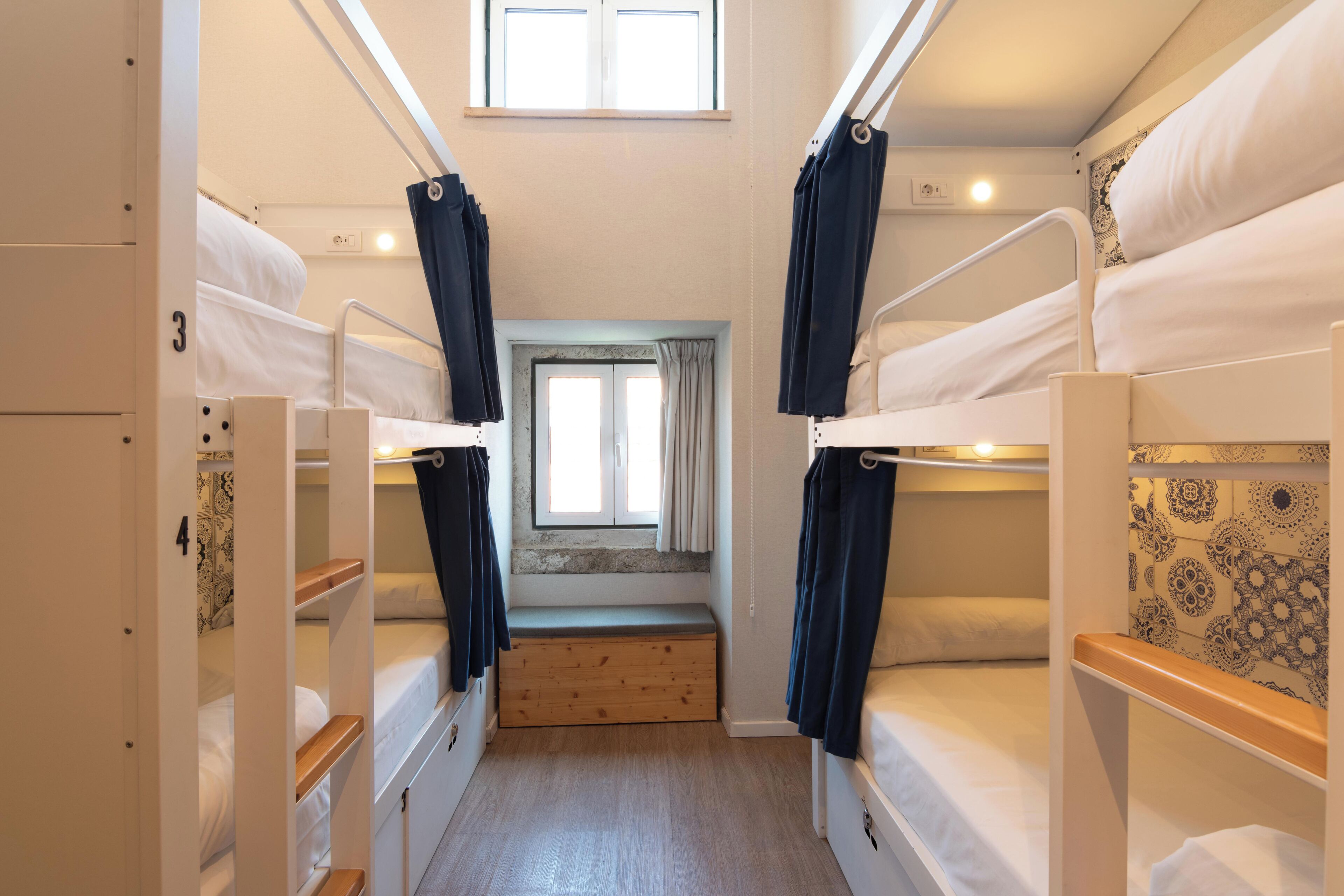 A narrow, well-lit room with bunk beds, a small window with curtains, and a wooden bench.