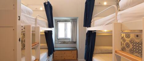 A narrow, well-lit room with bunk beds, a small window with curtains, and a wooden bench.