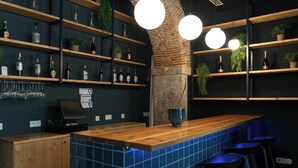 A bar area with wooden shelves holding bottles and glasses, a wooden counter, and blue-tiled backsplash.