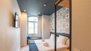 A hotel room with two beds, a bench, a window with curtains, patterned wallpaper, and a mounted TV.