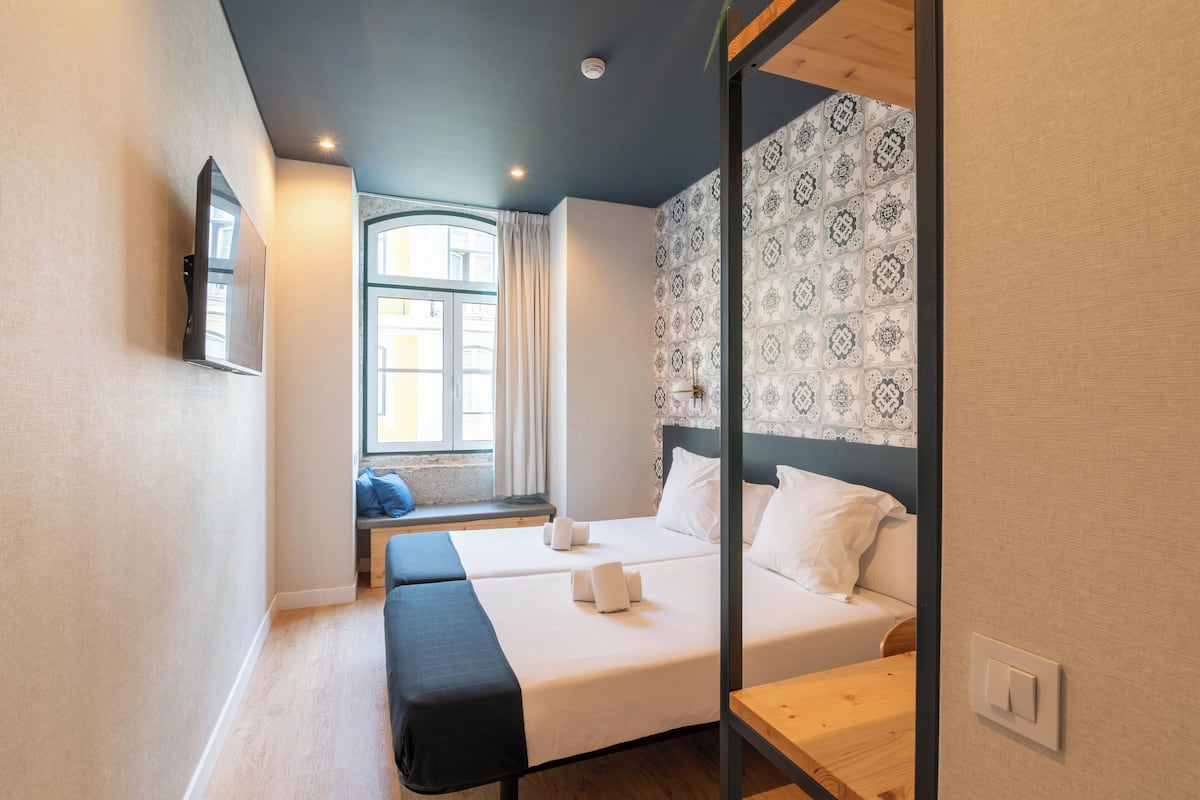 A hotel room with two beds, a bench, a window with curtains, patterned wallpaper, and a mounted TV.