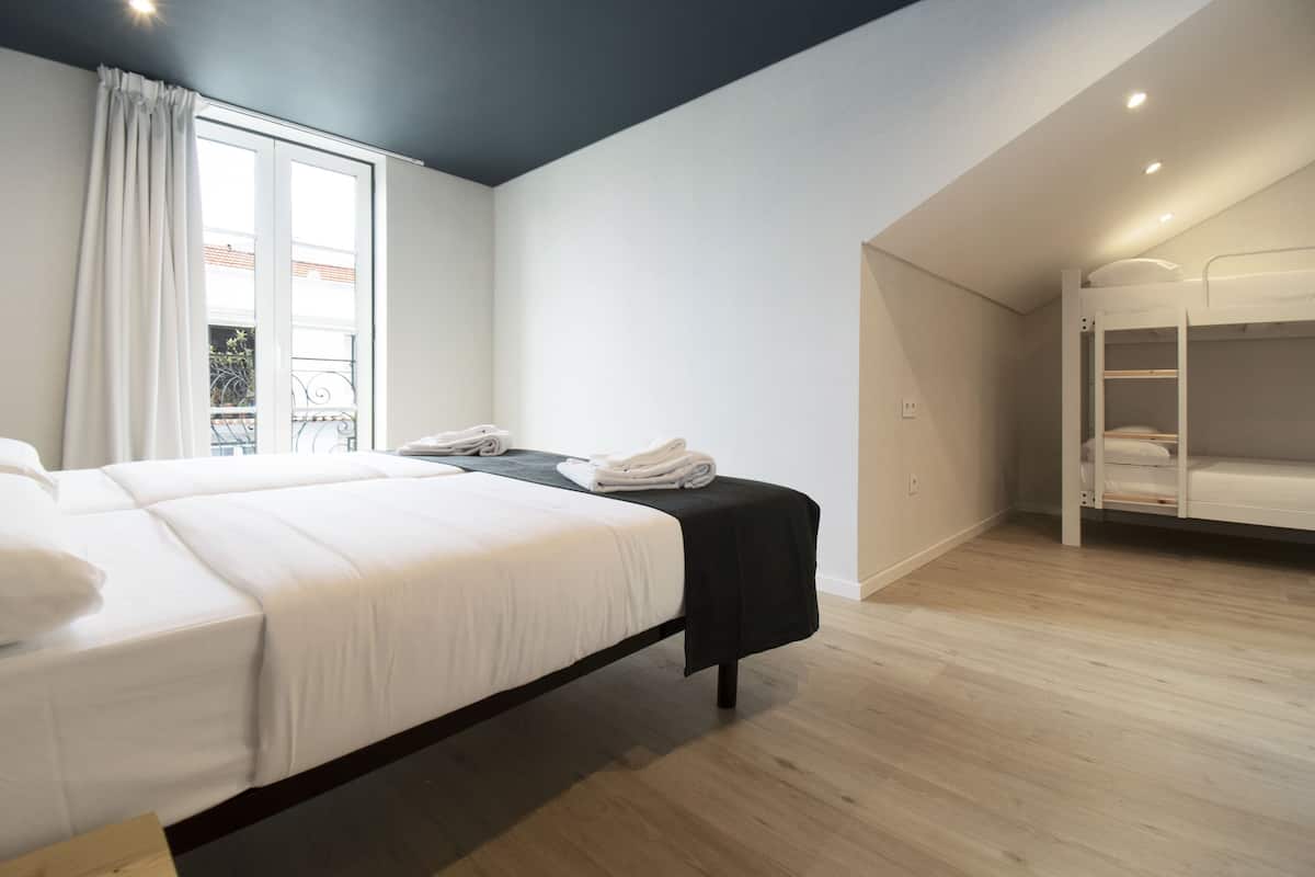 A modern hotel room with a large bed, a small table with a chair, a bunk bed, and a window with curtains.