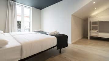 Una habitación de hotel moderna con una cama grande, una mesita con una silla, una cama litera y un ventanal con cortinas.
