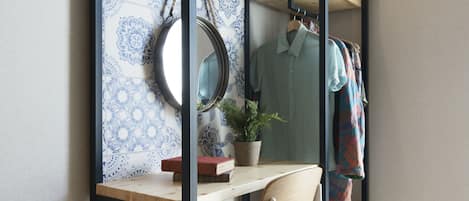 A room with a wooden desk, a mirror, a potted plant, and a clothing rack with hanging garments.