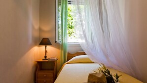 A bedroom with a bed, a nightstand, a lamp, a window with sheer curtains, and a straw hat on the bed. - Casa Paleopolis near Royal Baths MonRepo (Corfu)