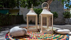 Two white lanterns on a colorful woven mat with rocks, set outdoors with trees and a building in the background. - Casa Paleopolis near Royal Baths MonRepo (Corfu)