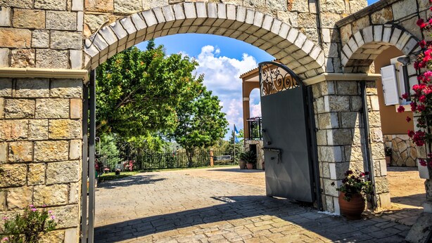 An arched stone gateway leading to a courtyard with a stone wall, potted plants, and a clear blue sky. - Hidden gem in nature reserve on the beach (Corfu)