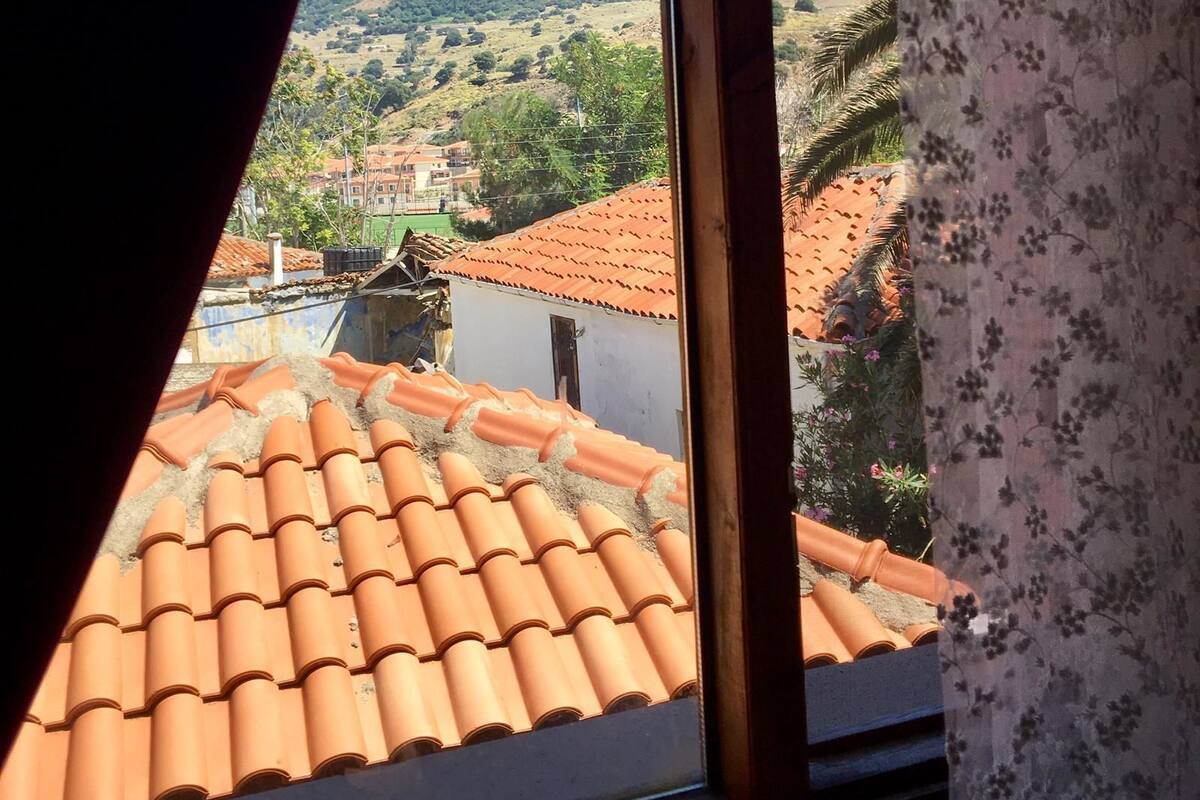 A view through a window with a floral curtain, revealing a hillside with terraced buildings and a red-tiled roof.