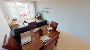 Uma sala de jantar com uma mesa de madeira posta para quatro pessoas, rodeada por cadeiras de couro. Um sofá e uma televisão estão visíveis ao fundo.