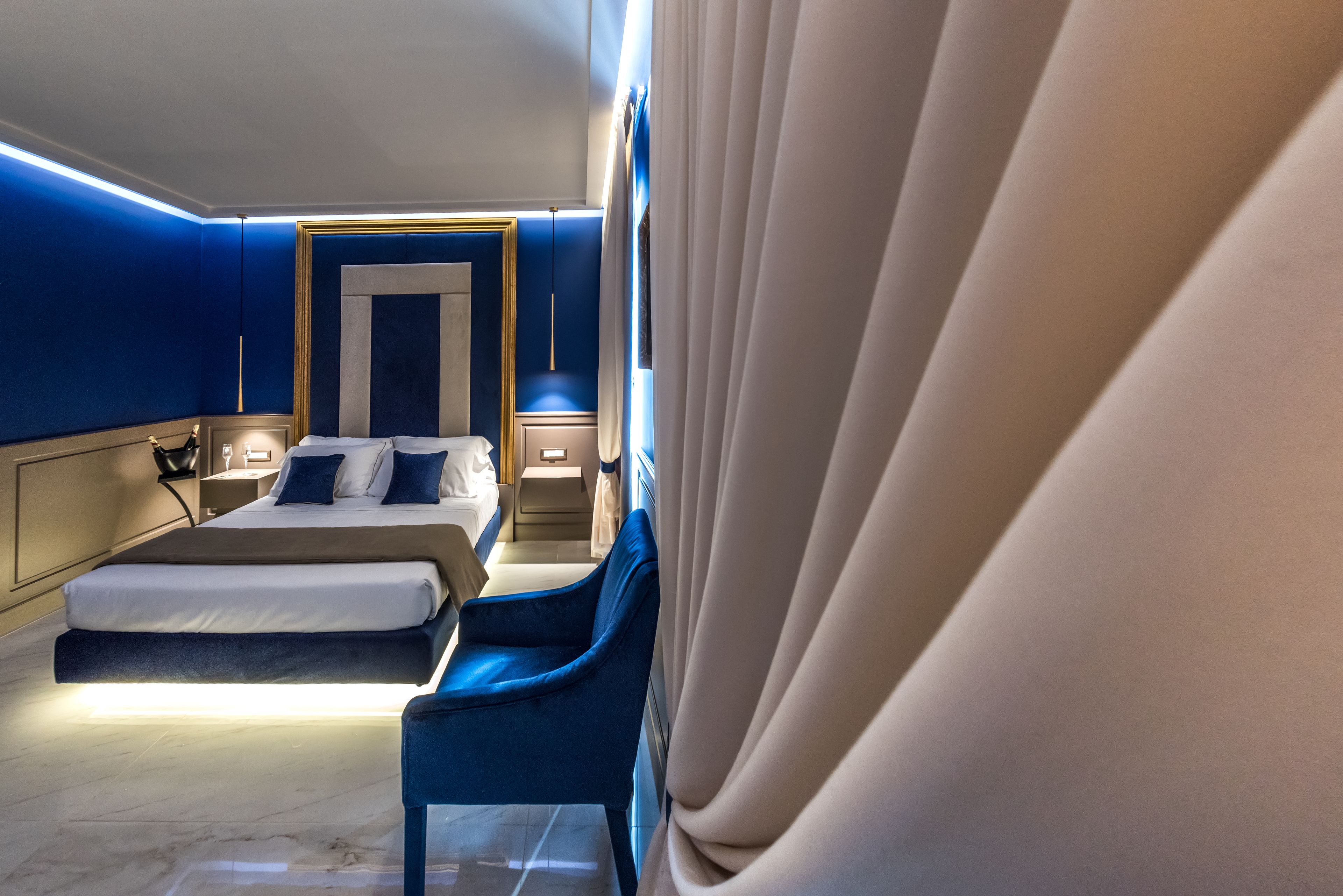 Una habitación de hotel moderna con una cama grande, un sillón azul y una mesita de noche con lámpara.