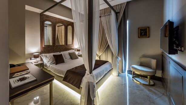 Un dormitorio con una cama con dosel, una mesita de noche con lámpara, una silla y un televisor.