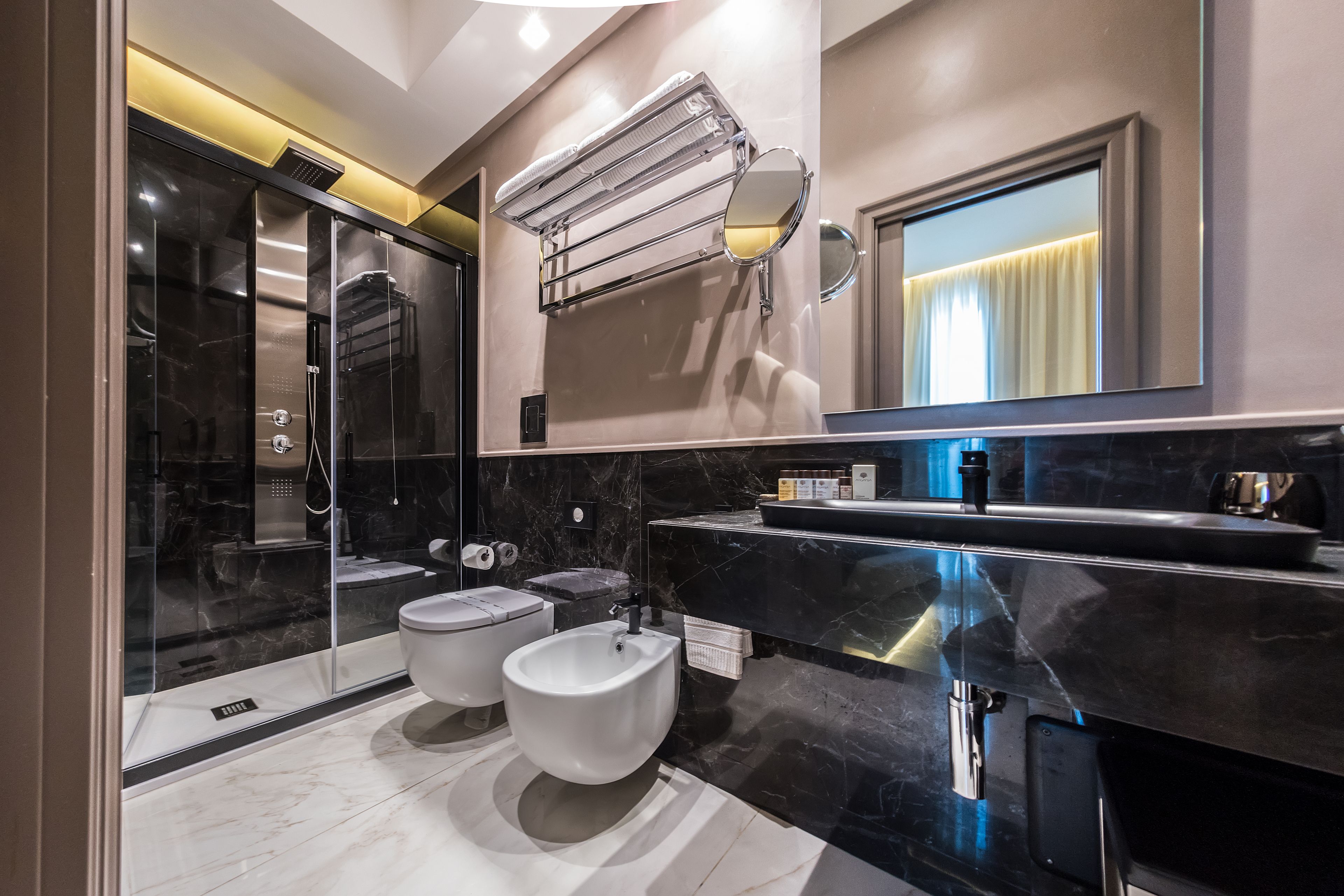 A modern bathroom with a glass-enclosed shower, two bidets, a large mirror, and a black marble countertop.