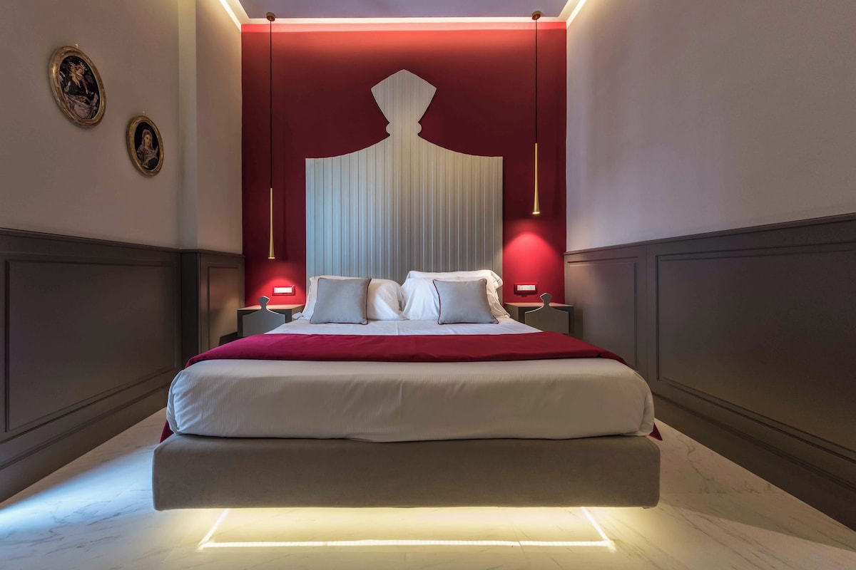 Un grand lit recouvert d’une couverture rouge, deux tables de chevet et des lampes fixées au mur.