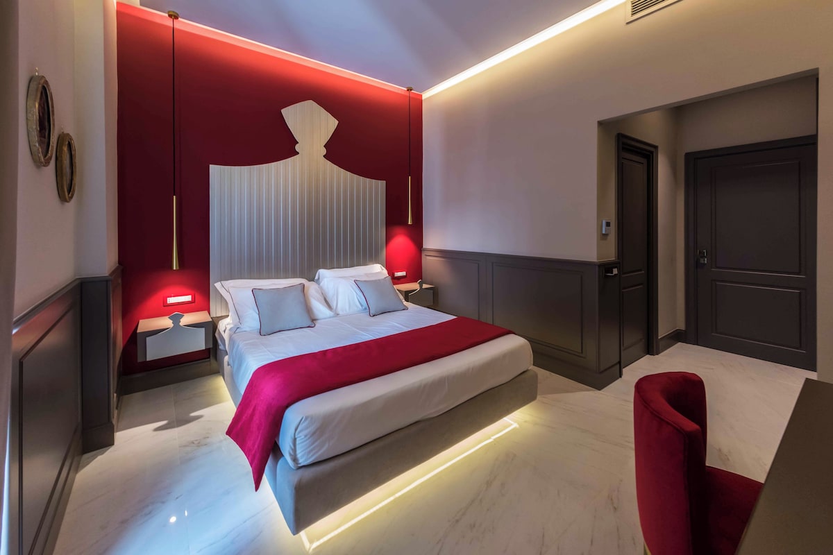 A modern bedroom with a large bed, bedside tables, a red accent wall, and a red chair.