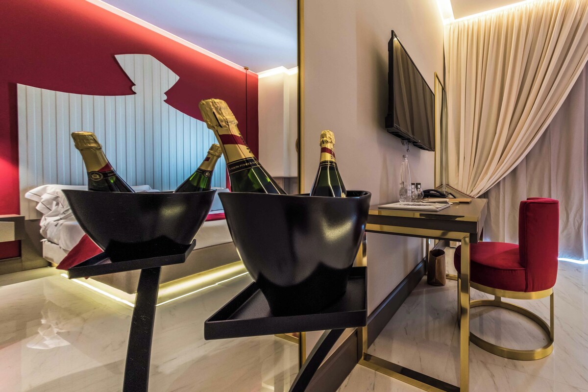 A modern hotel room with a bed, a desk with a computer, a TV, and two champagne buckets on a tray.