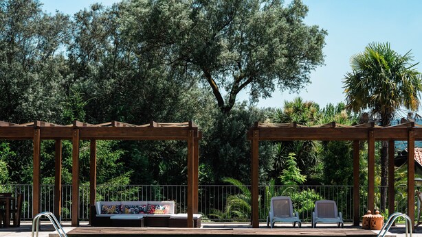 Un área de piscina con pérgolas de madera, sillones de descanso y un sofá con cojines de colores.
