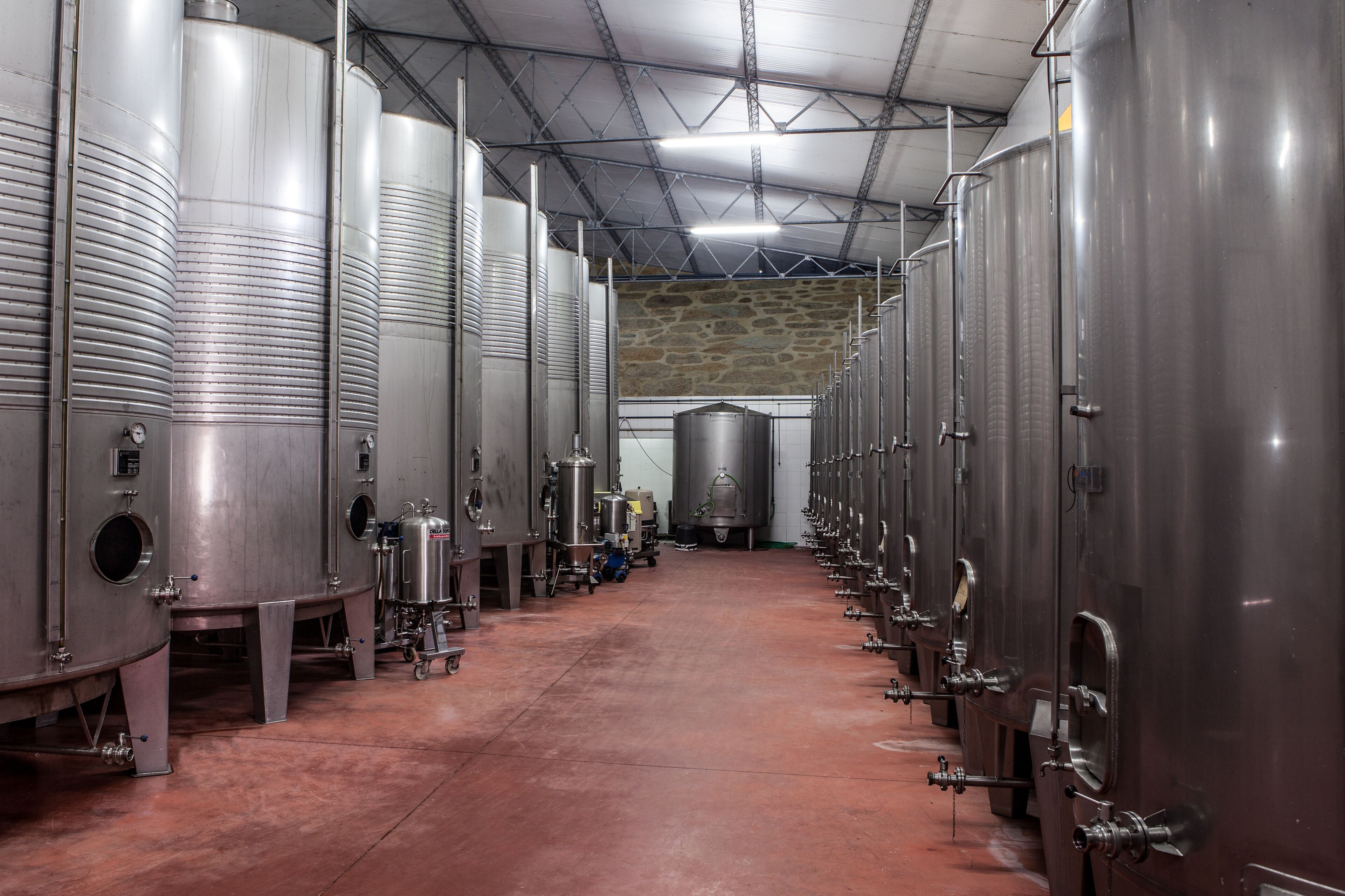 A row of stainless steel fermentation tanks in a brewery with a stone wall in the background.