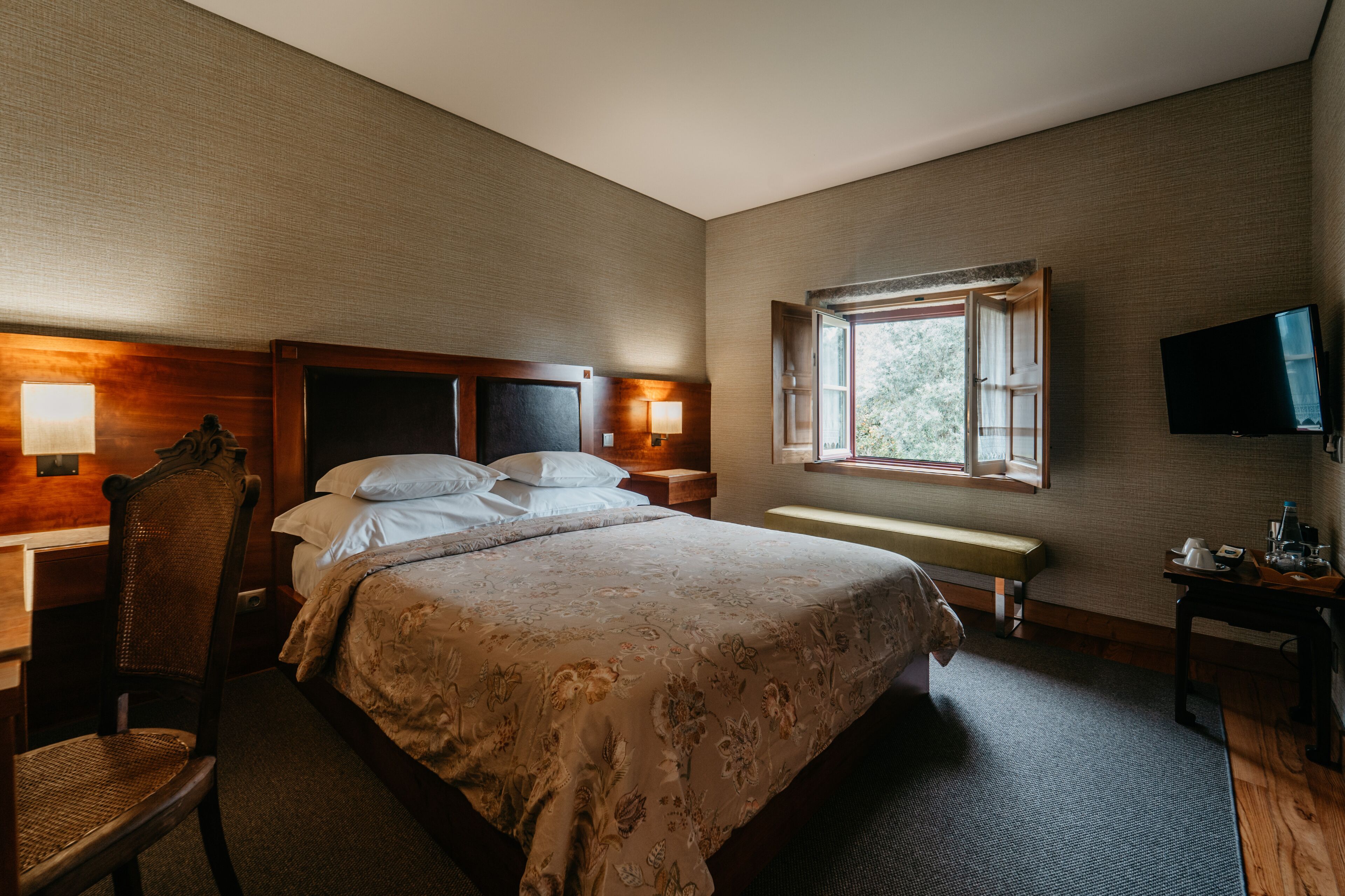 A hotel room with a large bed, bedside tables with lamps, a television, and a window with a view of trees.