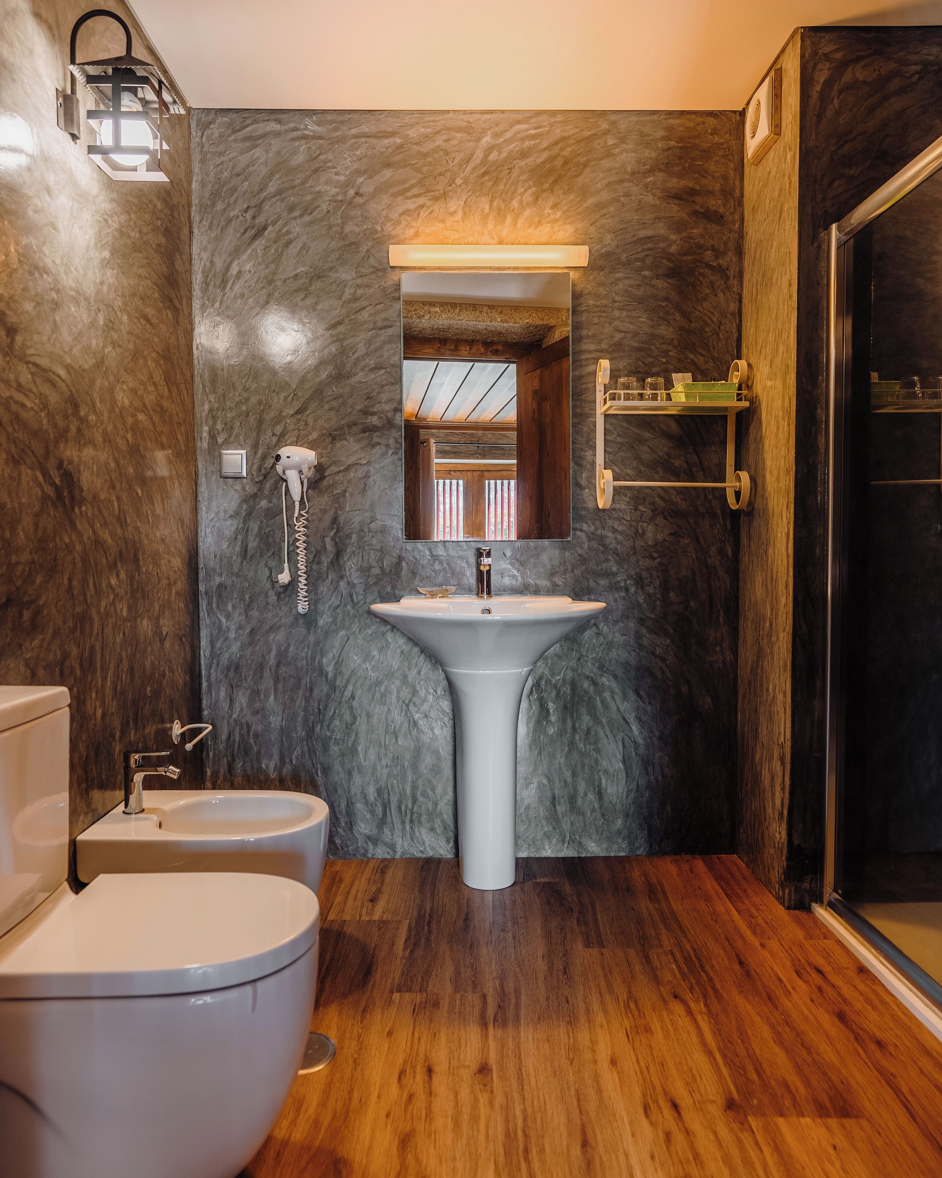 A bathroom with a white sink, a toilet, and a bidet. There is a mirror above the sink and a light fixture on the wall.