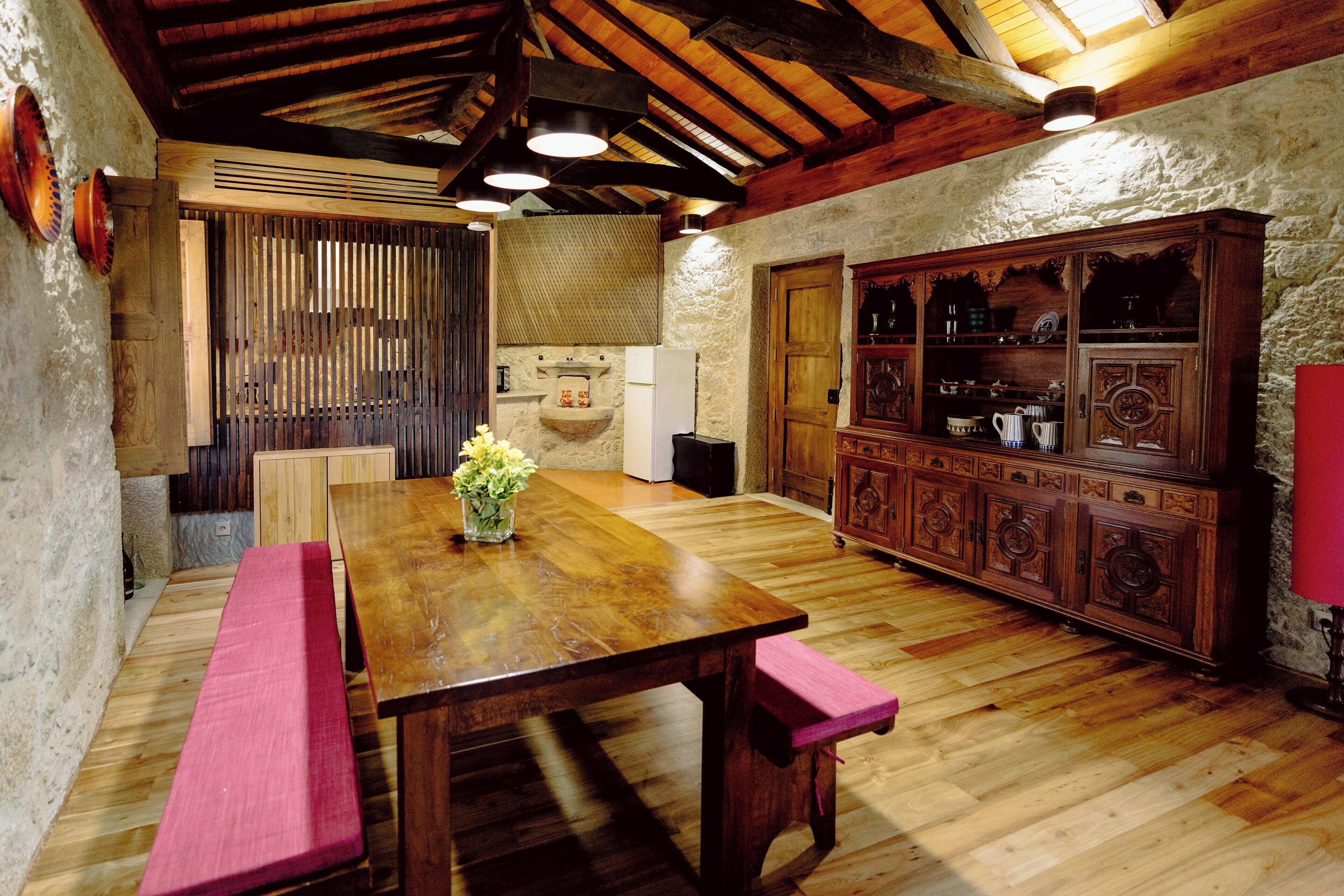 A rustic kitchen with wooden flooring, a large wooden table with pink cushions, and a cabinet with glass doors.
