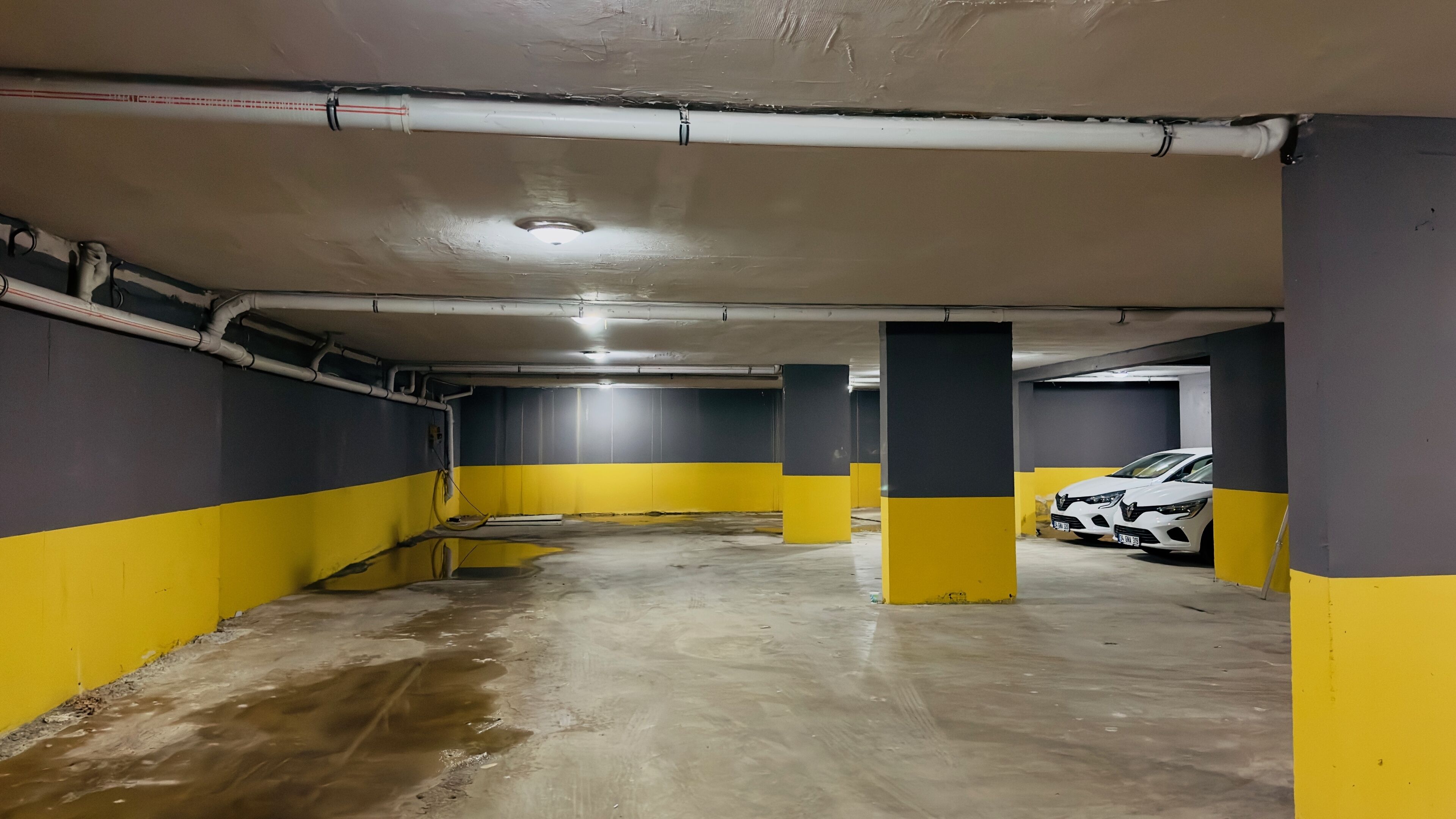 Un aparcamiento con paredes amarillas y grises, suelo de hormigón y coches blancos estacionados.
