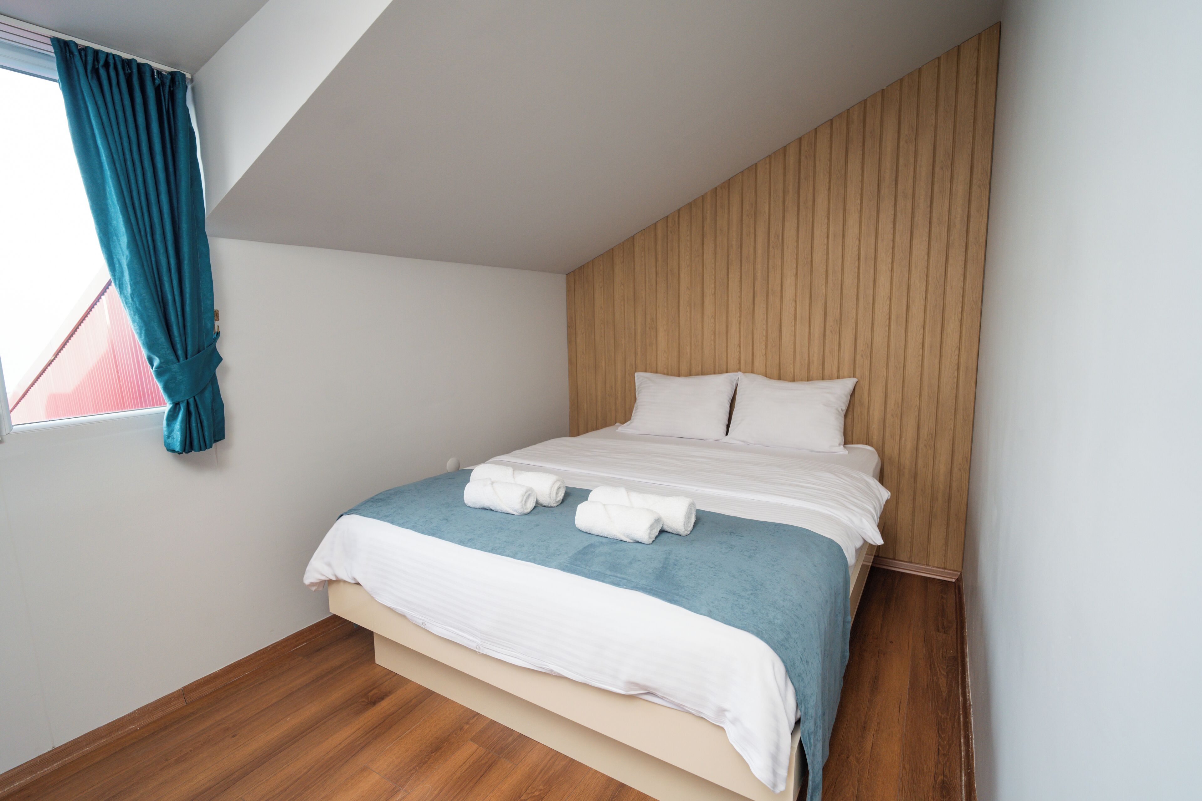 A neatly made bed with white and blue bedding, a wooden headboard, and a window with teal curtains.