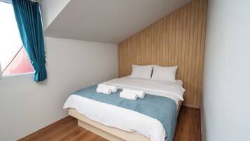 Una cama bien hecha con ropa de cama blanca y azul, un cabecero de madera y una ventana con cortinas color aguamarina.