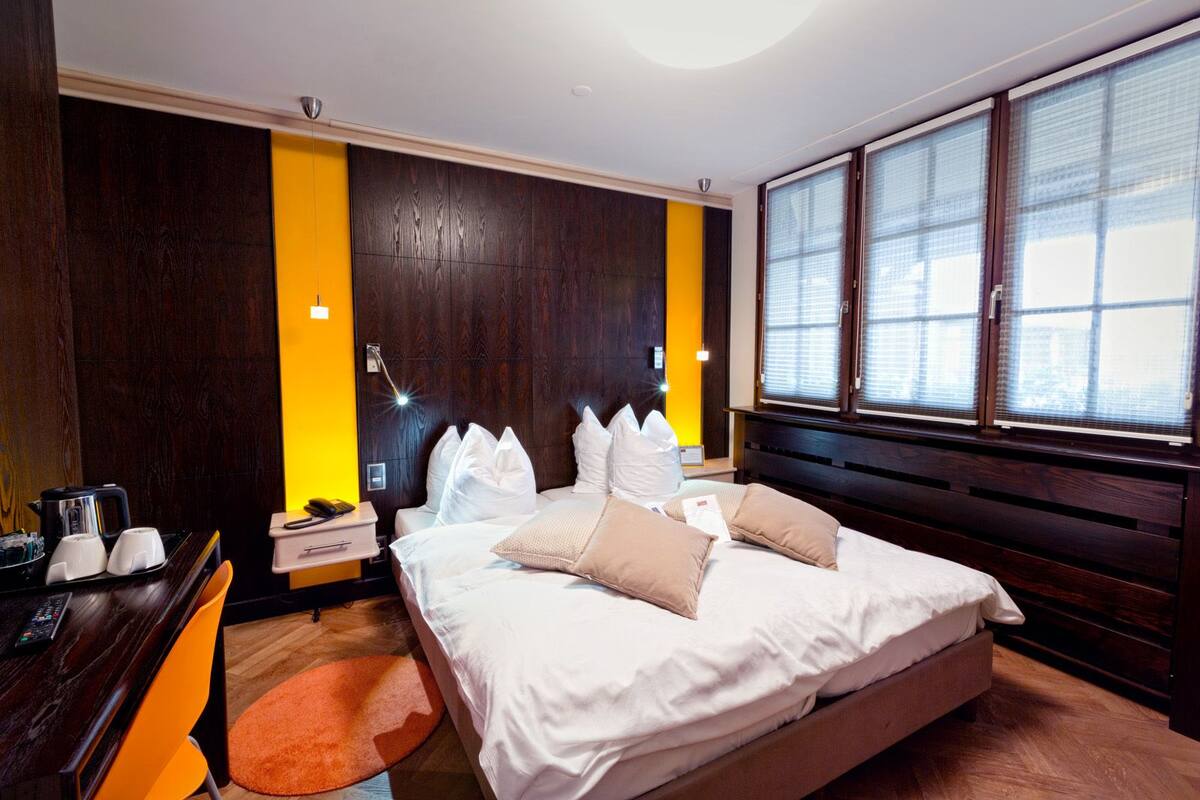 Habitación de hotel con una cama grande, un escritorio con silla, una mesita de noche y una ventana con persianas.