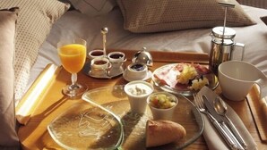 A neatly made breakfast tray on a bed with a wooden headboard, including a glass of orange juice, various condiments, a croissant, and a fruit bowl. - Hotel Restaurant & Spa - Le Cerf (Marlenheim)