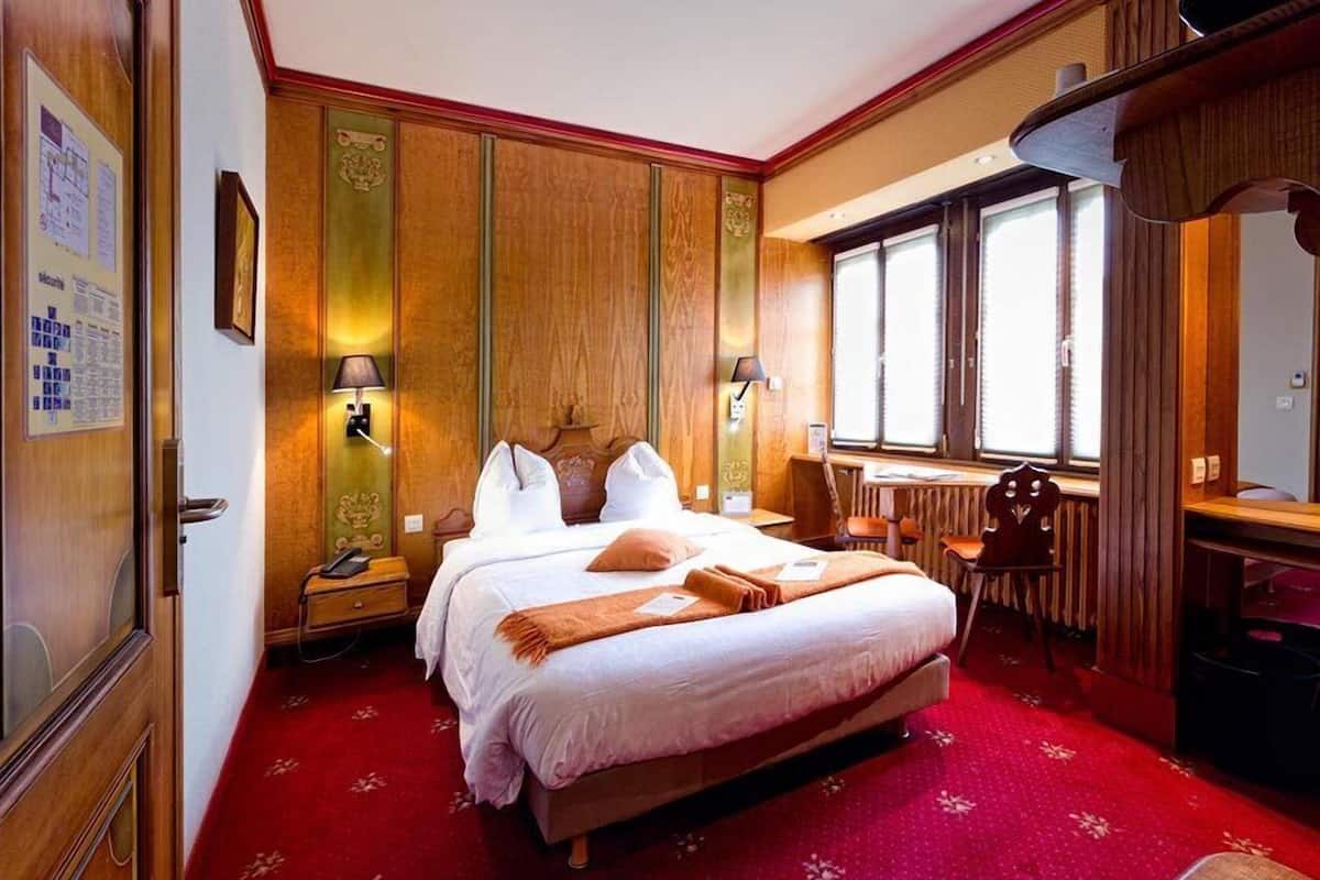 Habitación de hotel con una cama grande, dos mesitas de noche, un escritorio y una silla. Hay dos lámparas en la pared y una ventana con cortinas.