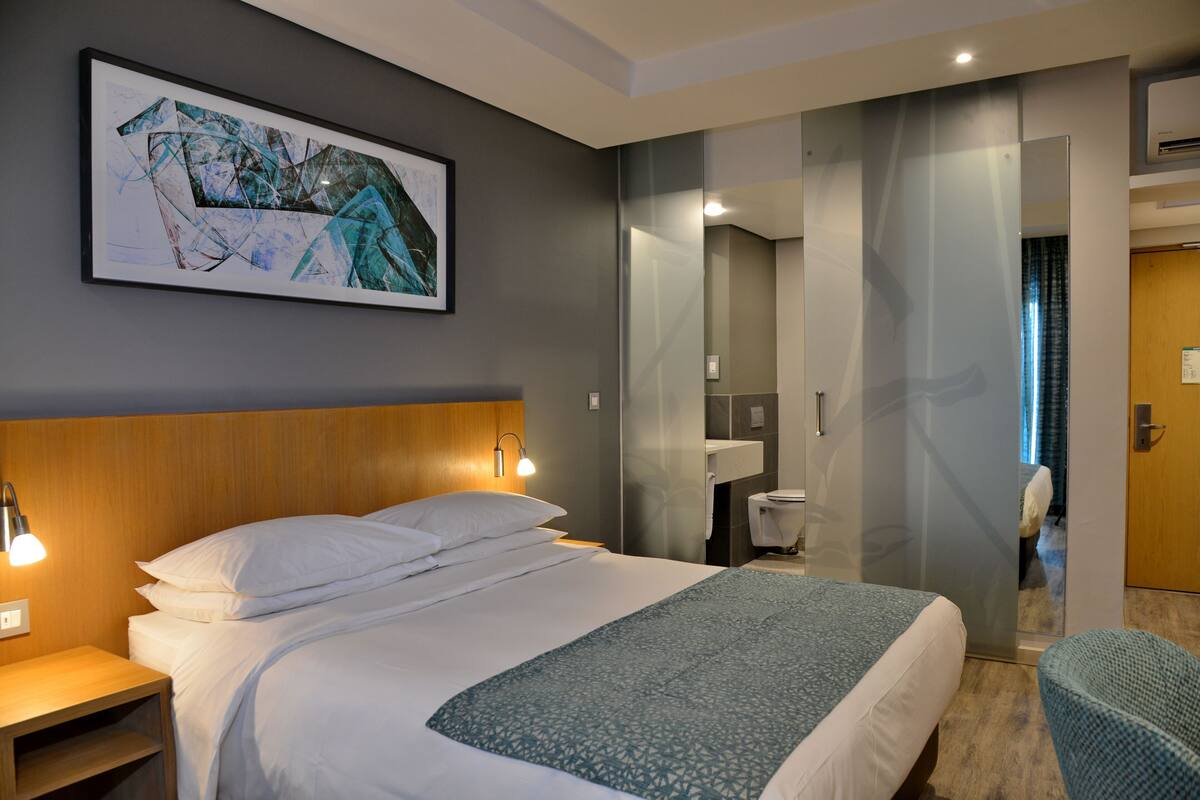 Ein Hotelzimmer mit einem großen Bett, einem Nachttisch, einem Wandbild, einem Spiegel und Blick ins Bad.