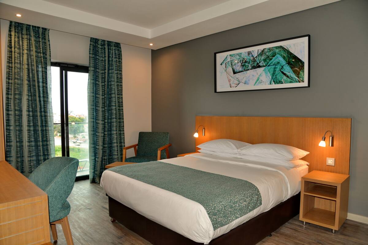 Ein Hotelzimmer mit einem großen Bett, einem Sessel, einem Schreibtisch und Blick ins Freie durch eine Glastür.