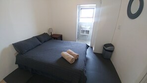 A bedroom with a large bed, a bedside table with a lamp, a small trash bin, and a window with a view of the outside. - Blouberg Beach Retreat (Cape Town)