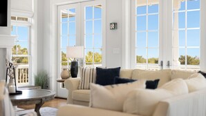 Un salon spacieux avec un grand canapé beige, une petite table ronde avec une lampe et une vue sur l’extérieur grâce à de grandes fenêtres.