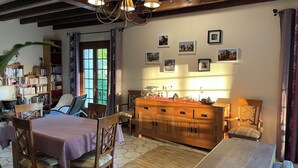 A cozy room with a wooden dining table, chairs, a sofa, a bookshelf, and a large window with curtains. - Au Pre des Sources (Campagne)