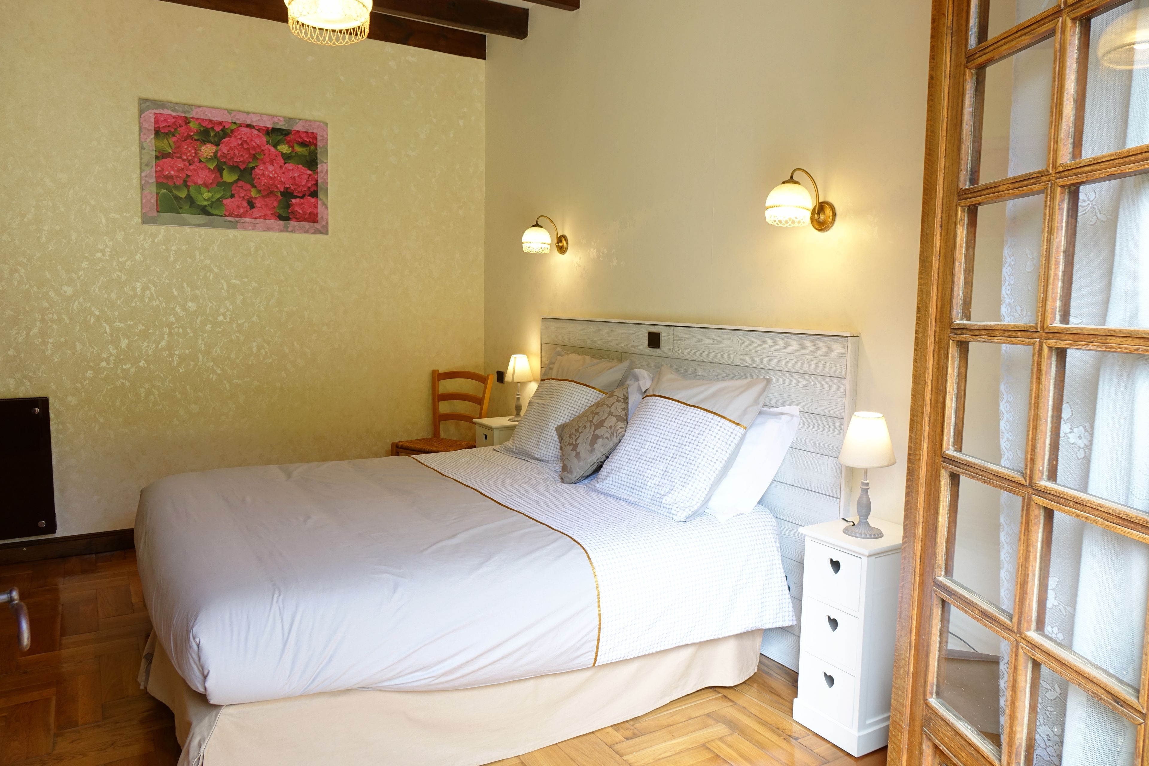 Una cama bien hecha con colcha de cuadros blancos y azules, dos mesitas de noche con lámparas, una silla de madera y una ventana con cristal satinado.