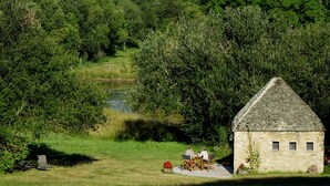 A stone building with a triangular roof, surrounded by lush greenery and a small pond. - Au Pre des Sources (Campagne)