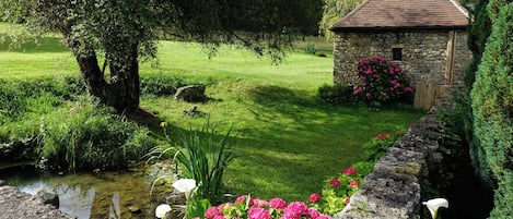 En bygning i sten med et tegltag, omgivet af en frodig have med et bassin og lyserøde blomster.