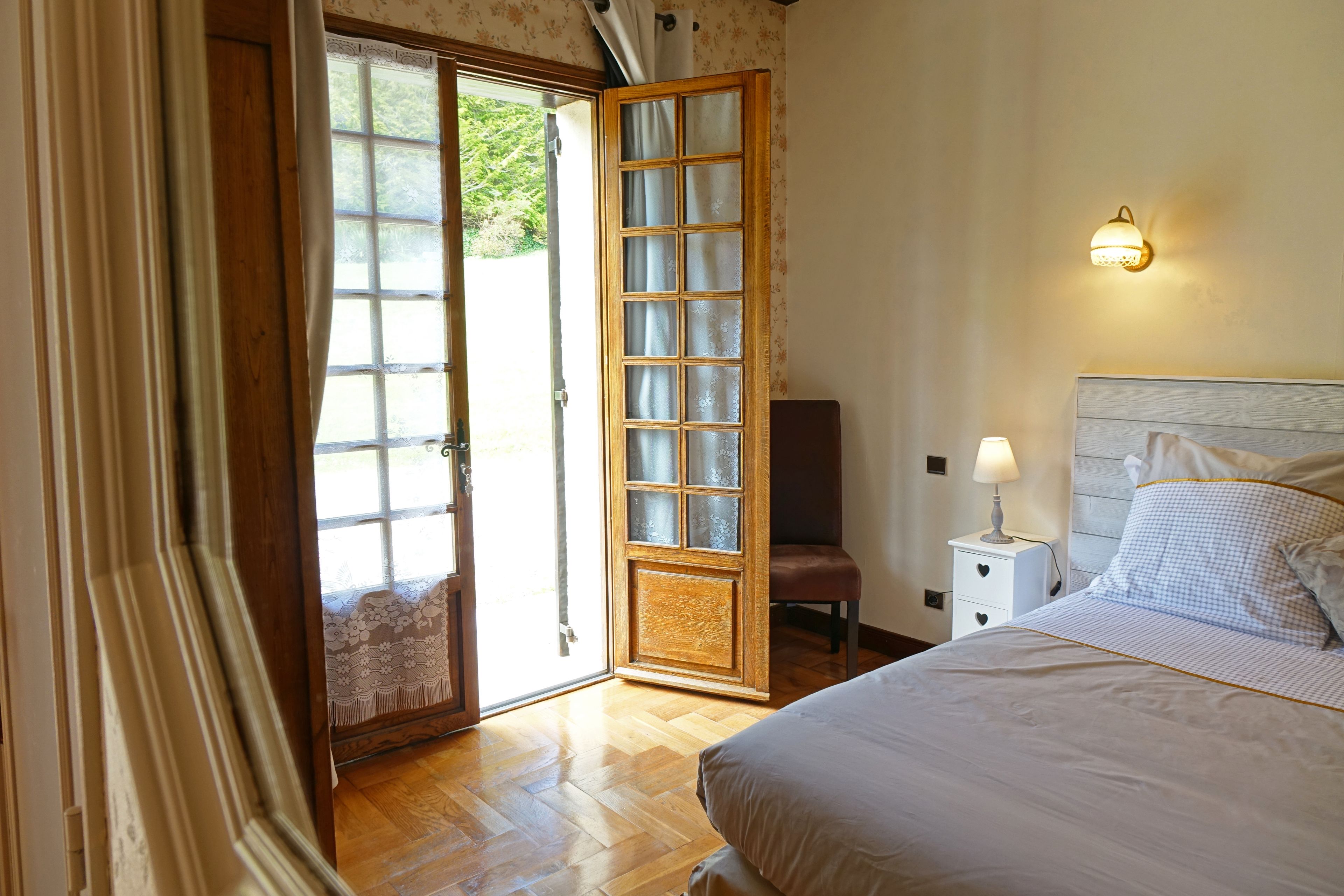 Een slaapkamer met houten vloer, een bed met wit bedlinnen, een nachtkastje met lamp en een raam met uitzicht op groen.