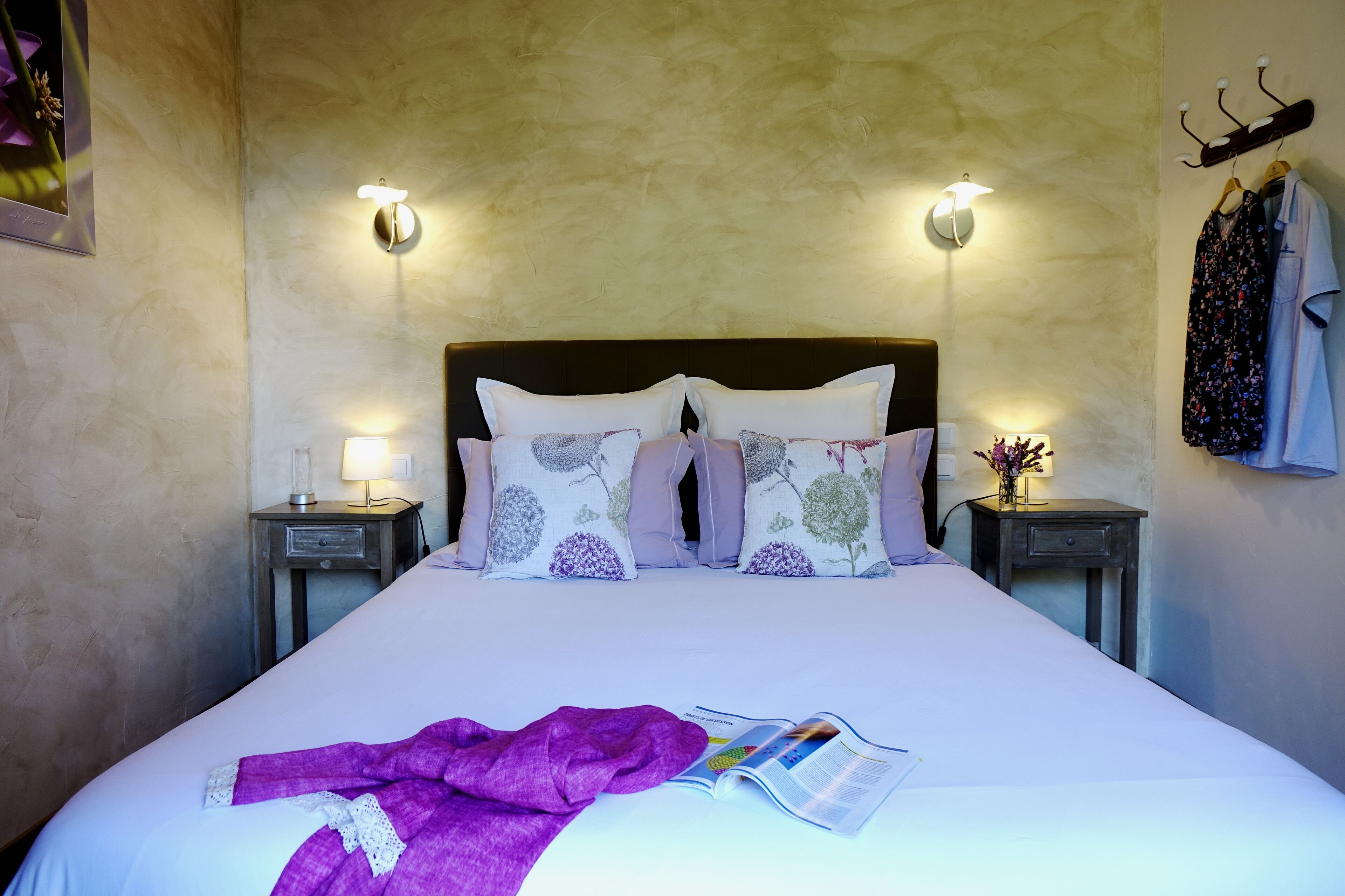 Un lit bien fait, recouvert d’une couverture et de coussins violets, deux lampes de chevet, une table de chevet avec une lampe, et un porte-manteau mural sur lequel est accroché un peignoir.