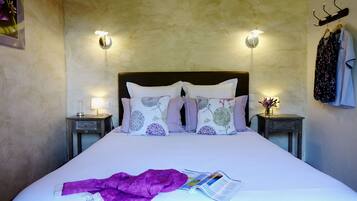 Un lit bien fait, recouvert d’une couverture et de coussins violets, deux lampes de chevet, une table de chevet avec une lampe, et un porte-manteau mural sur lequel est accroché un peignoir.