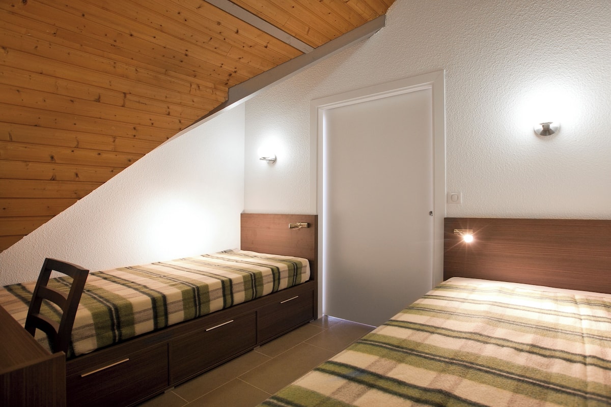 A bedroom with a sloped ceiling, a bed with a plaid bedspread, a nightstand, and a door.