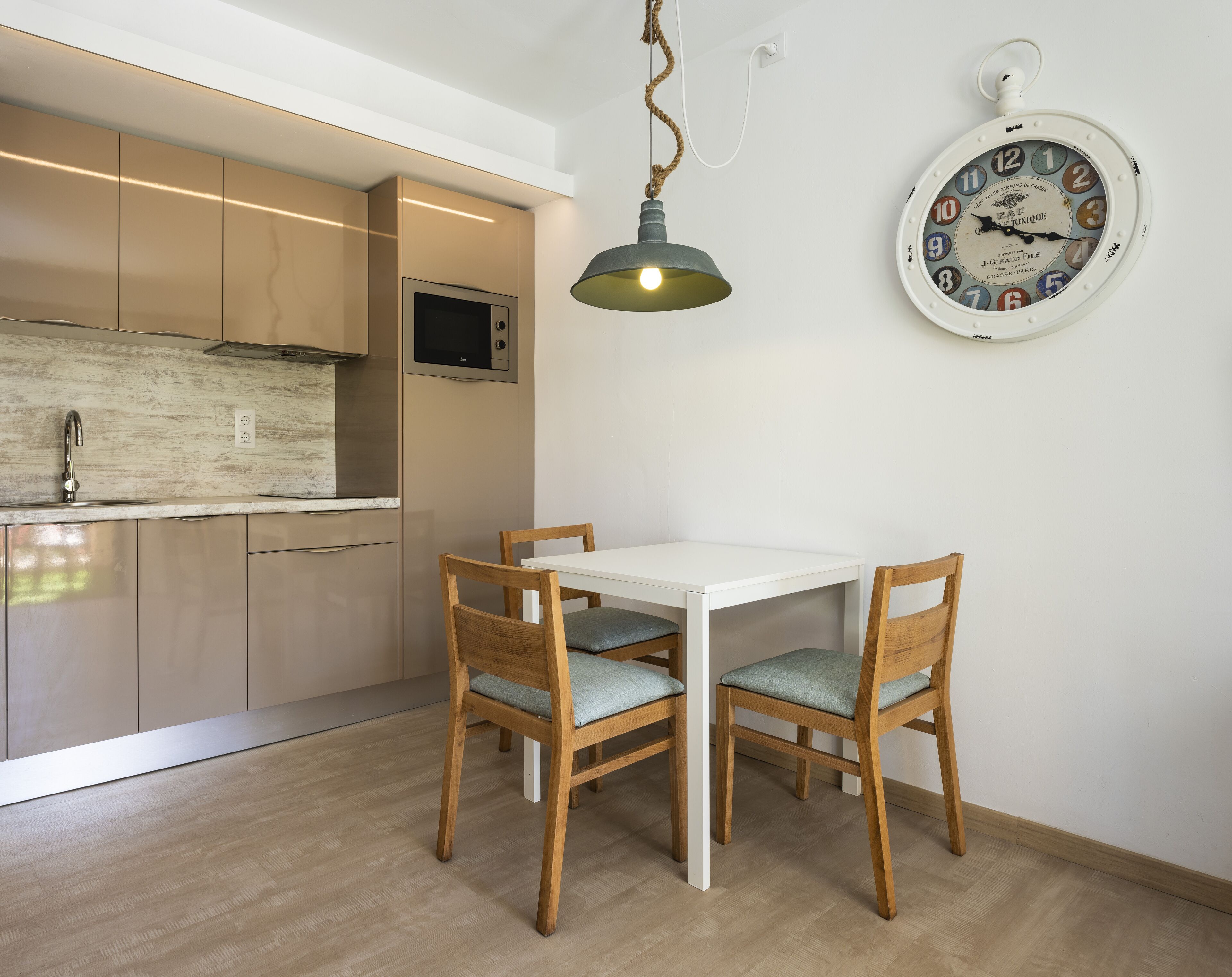 A modern kitchen with wooden cabinets, a white dining table, and two wooden chairs with grey cushions. A vintage-style wall clock is mounted on the wall.