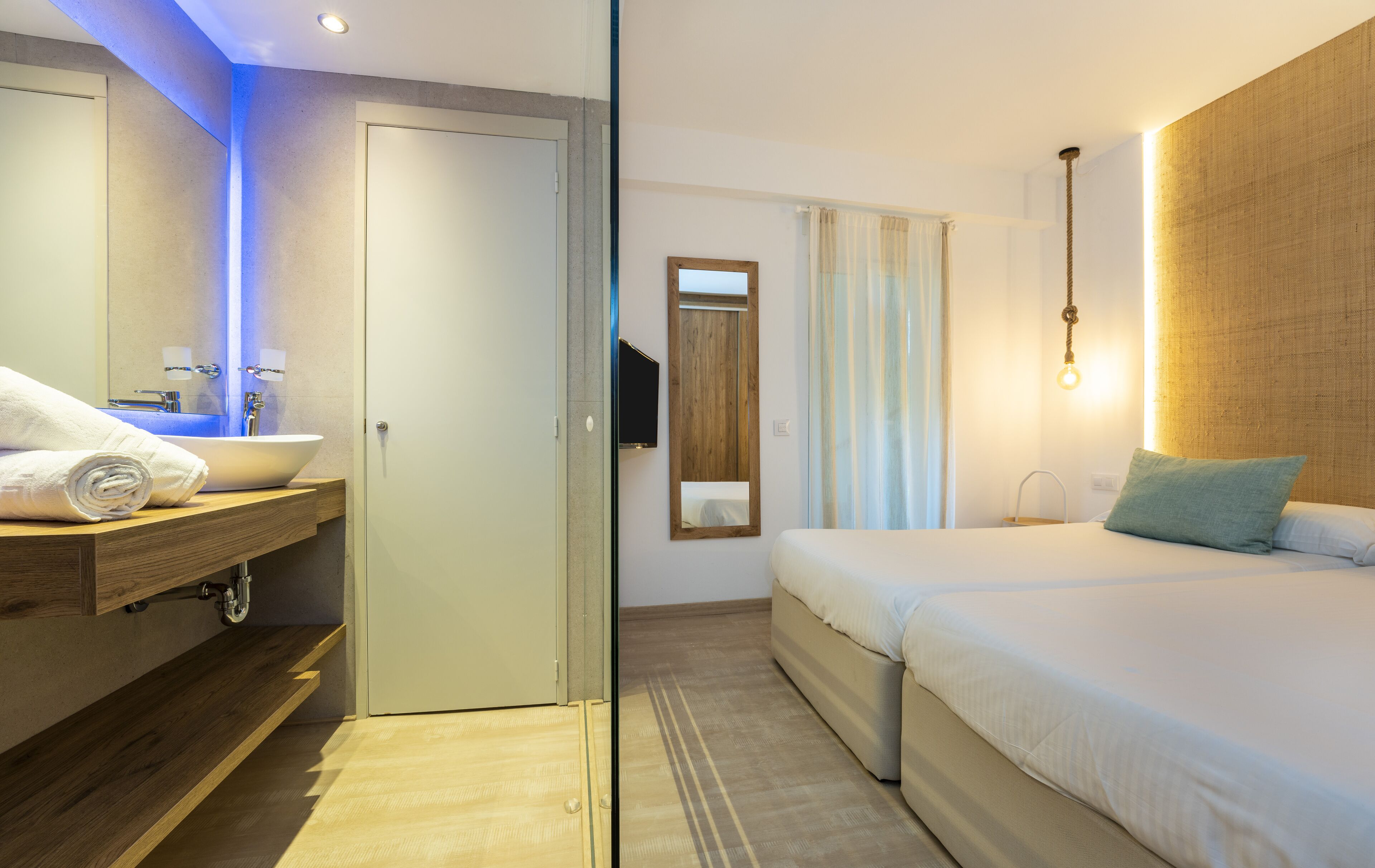 A hotel room with a bathroom, a bed, a wooden shelf, a mirror, and a bedside table.