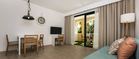 A modern hotel room with a wooden dining table, two chairs, a TV, a sofa with cushions, a lamp, and a sliding glass door leading to an outdoor area.