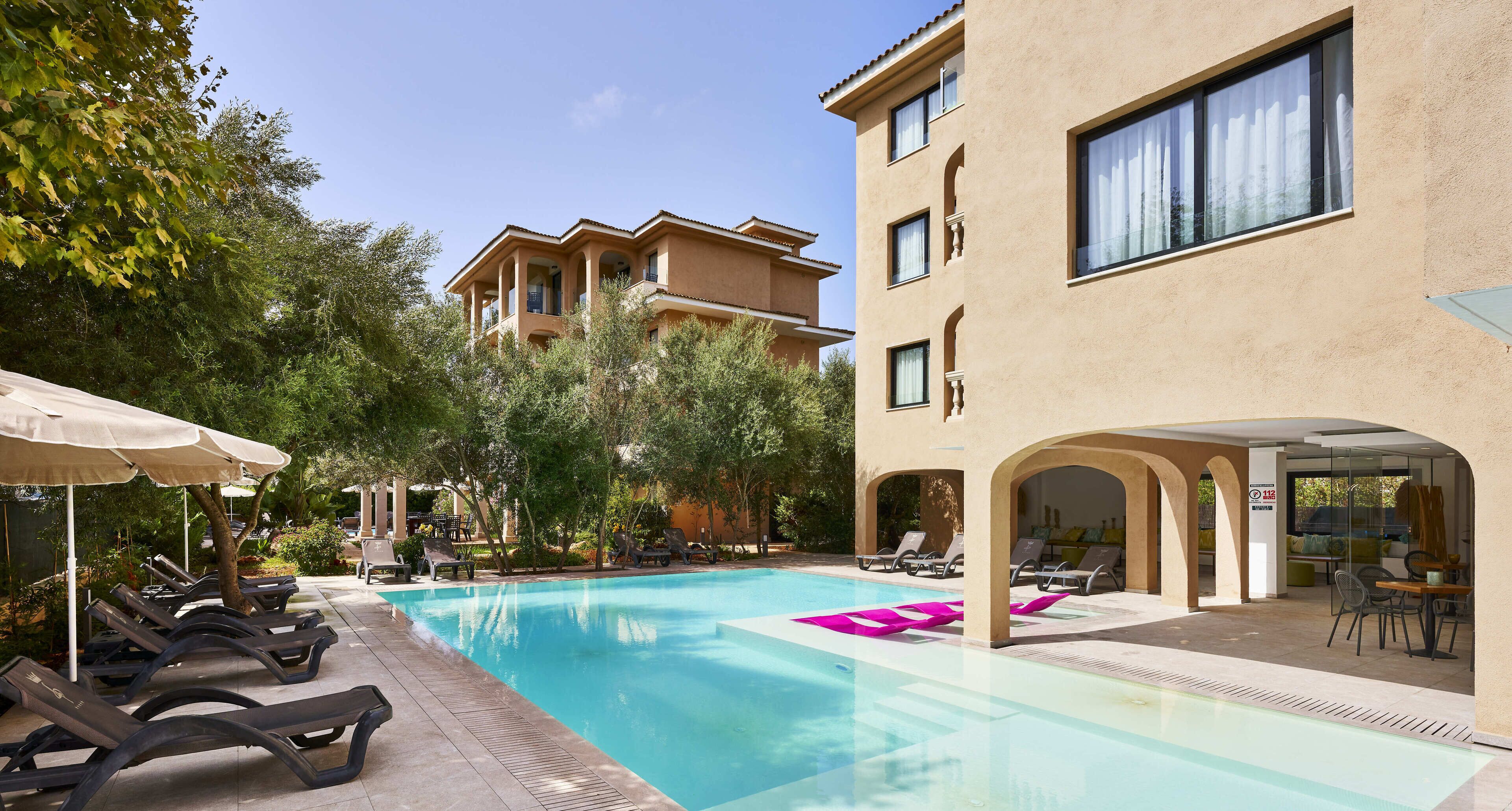 Un espace piscine avec des chaises longues et des parasols, un bâtiment à la façade beige et un ciel d’un bleu limpide.
