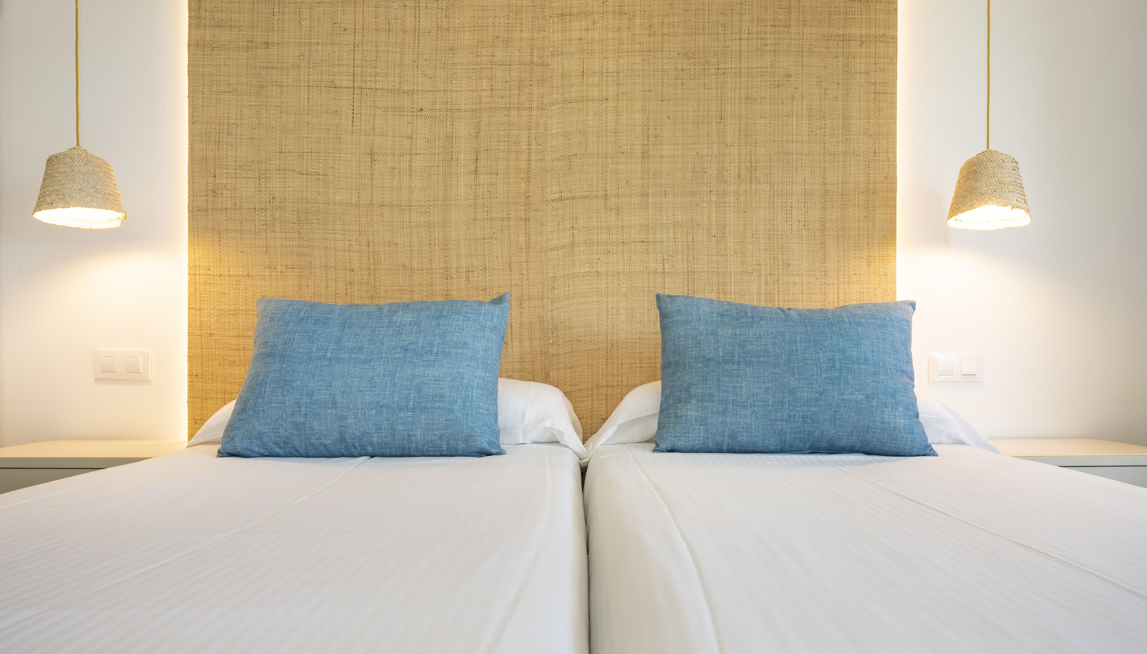 A double bed with white linens and two blue pillows, a woven headboard, and two hanging pendant lights.