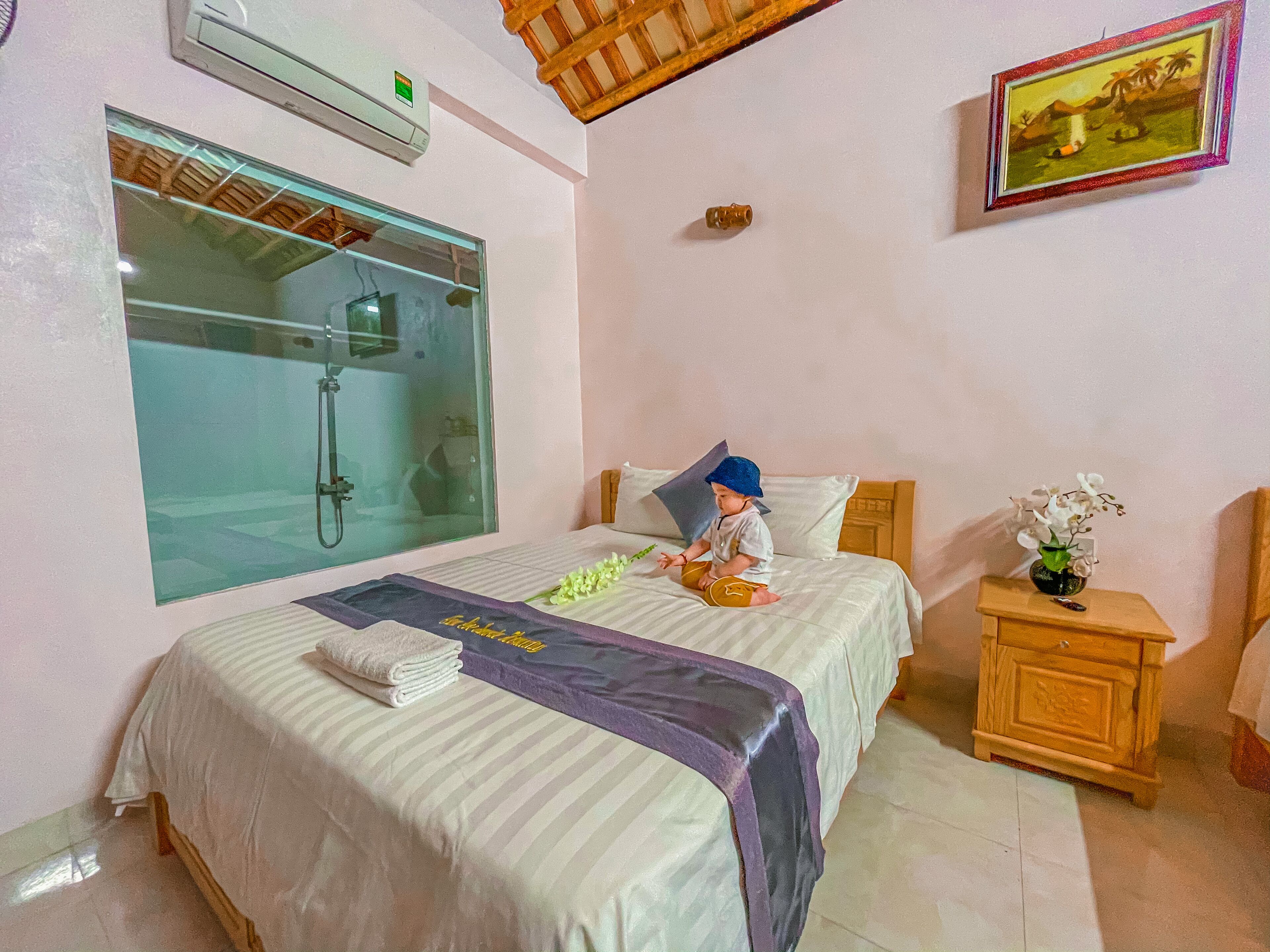 A hotel room with a bed, a nightstand, a window with a shower, and a painting on the wall.