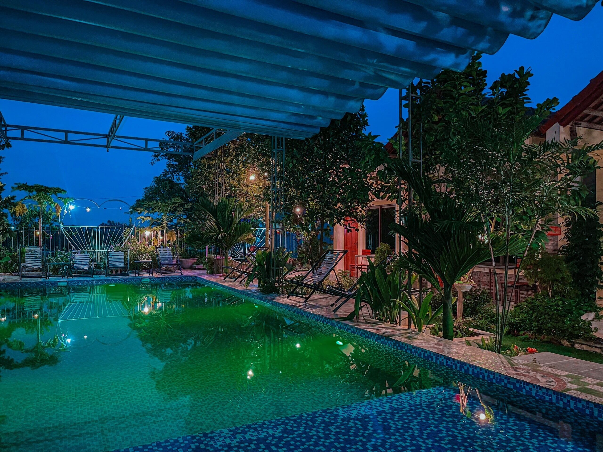 A nighttime pool area with a covered seating area, surrounded by lush greenery and a building with a red door.