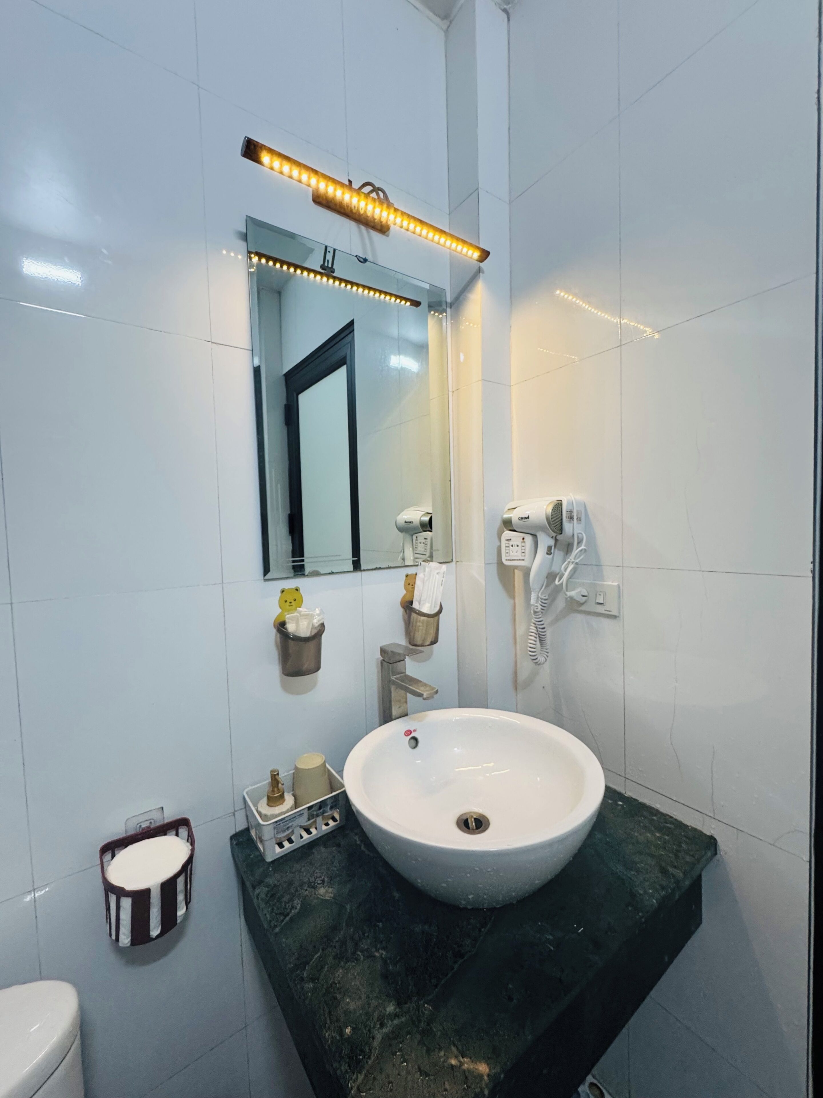 Uma casa de banho com uma pia branca, um espelho, um secador de cabelo e um porta-toalhas.